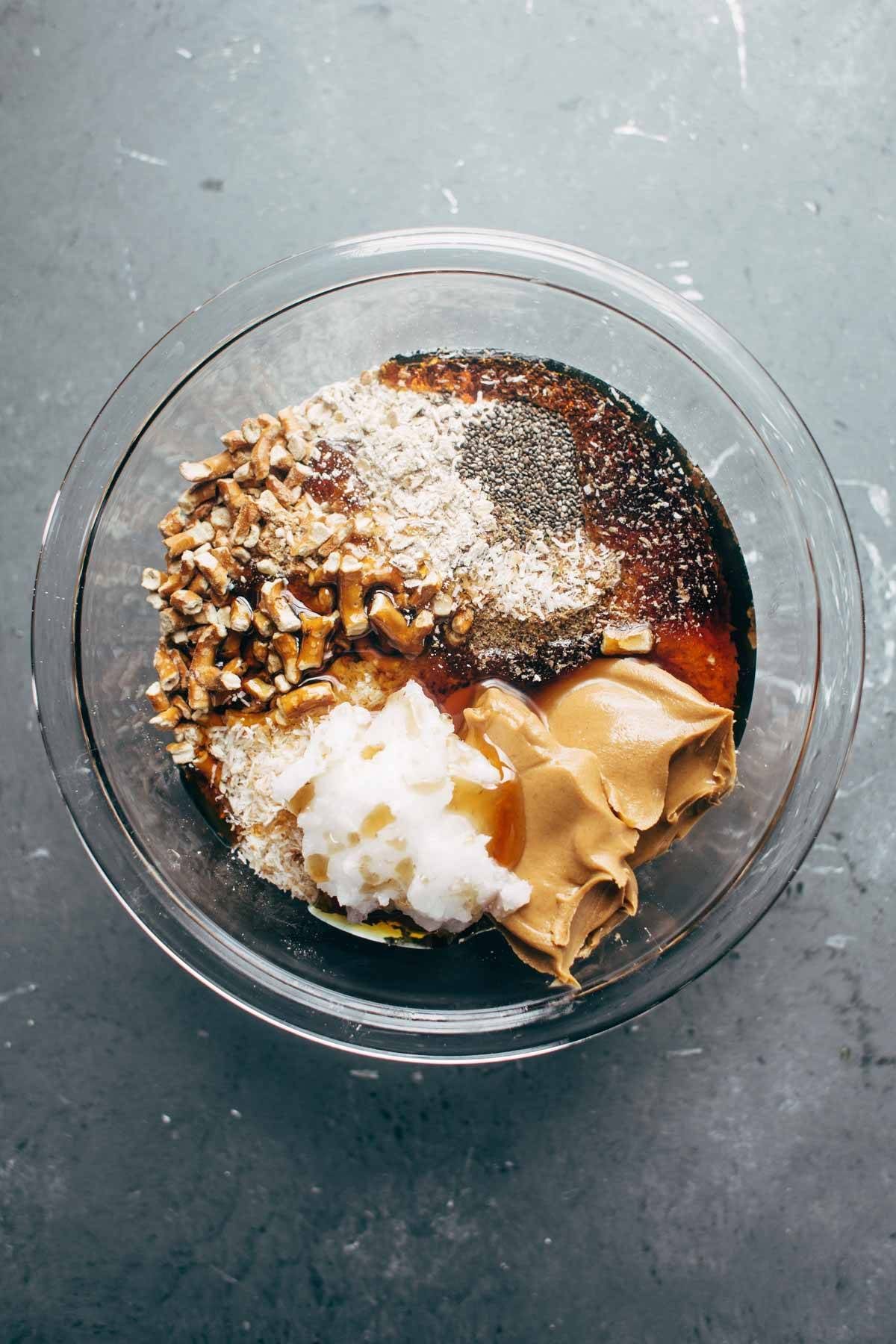 Pretzels, coconut oil, seeds, syrup, and peanut butter in a clear bowl.
