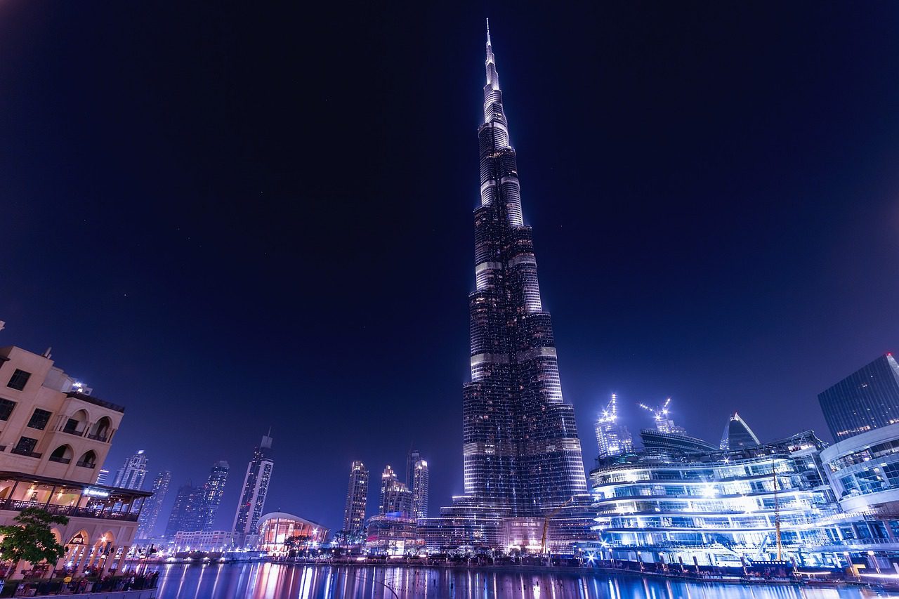 A nighttime view of Dubai's iconic Burj Khalifa, glowing brightly against the dark sky, surrounded by modern architecture and serene reflections on the water.